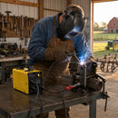  Welder in leather apron using yellow Mini Welding Machine on metal part in rustic farm workshop